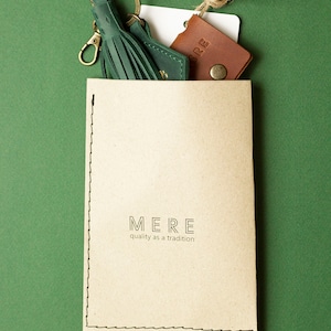 May include: A green leather tassel keyring with a gold-coloured clasp and ring, a small brown leather cardholder, and a paper card with the word "MERE" and the phrase "quality as a tradition" printed on it. The items are on a green background.