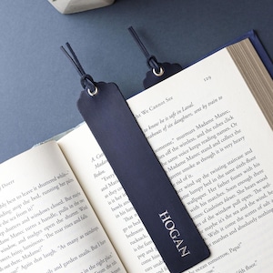 Op de afbeelding: Marineblauw leren bladwijzer met de naam "HOGAN" in witte letters. De bladwijzer is in een boek geplaatst. Een tweede bladwijzer is zichtbaar. Het boek heeft witte pagina's en een donkerblauwe kaft.