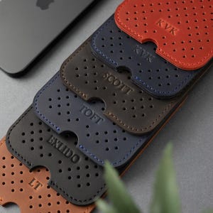 May include: A stack of personalized leather phone cases in shades of brown, navy, and red. Each case features a perforated design and is embossed with initials or a name. A gray smartphone with the Apple logo is visible in the background.