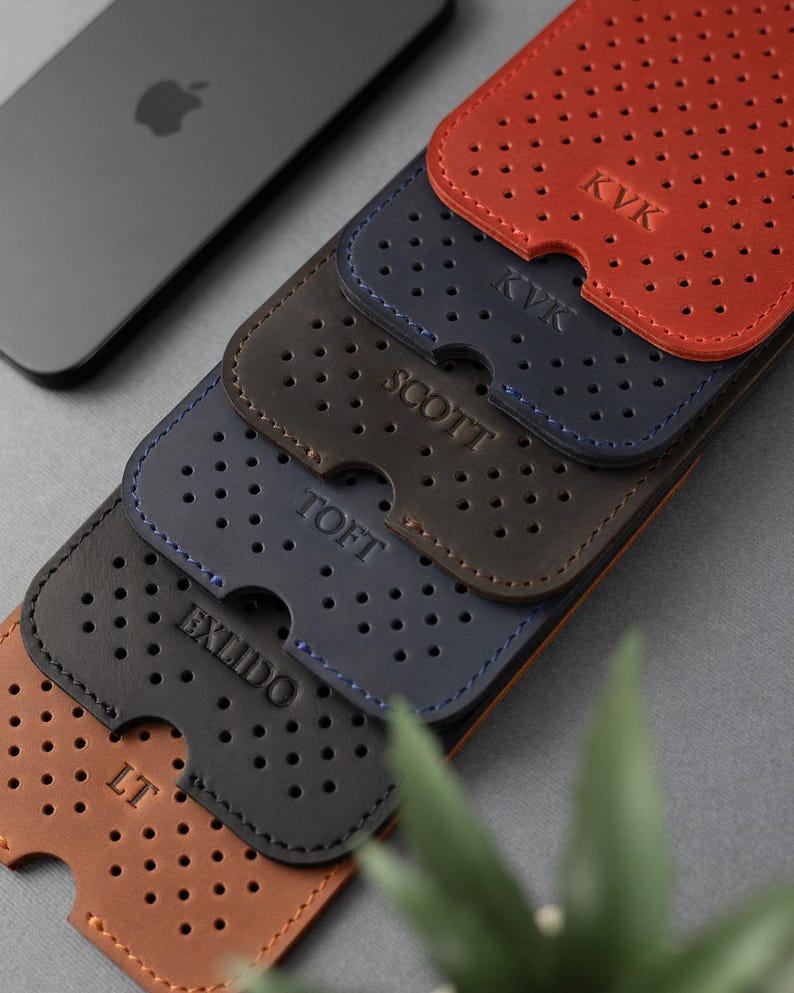 May include: A stack of personalized leather phone cases in shades of brown, navy, and red. Each case features a perforated design and is embossed with initials or a name. A gray smartphone with the Apple logo is visible in the background.