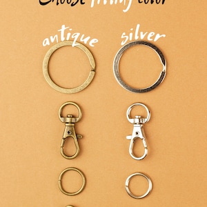 May include: A selection of keychains and hardware in antique brass and silver finishes. The image shows key rings, lobster clasps, split rings, and rivets. Text at the top reads "Choose fitting colour" with the options "antique" and "silver" below.