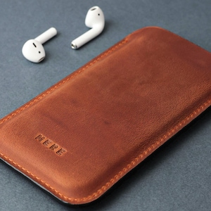 May include: A brown leather phone case with the word "MERE" embossed on the front. Two white wireless earbuds are in the background.