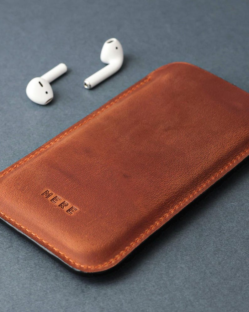 May include: A brown leather phone case with the word "MERE" embossed on the front. Two white wireless earbuds are in the background.
