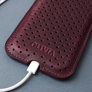 May include: A burgundy leather phone case with a perforated design and red stitching. The word "OLIVIA" is embossed on the case. A white charging cable is plugged into the bottom of the case.