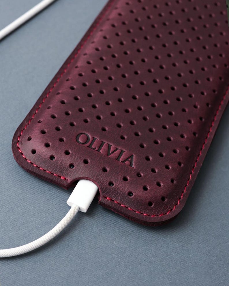 May include: A burgundy leather phone case with a perforated design and red stitching. The word "OLIVIA" is embossed on the case. A white charging cable is plugged into the bottom of the case.
