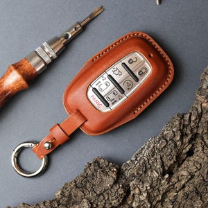 May include: A brown leather key fob cover with silver buttons and a metal keyring. The cover has the word "PANIC" on it. A leather strap connects the cover to the keyring. A screwdriver and a screw are also in the image.