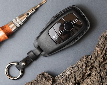 May include: A black leather car key fob cover with visible stitching, featuring a matching leather strap and a silver-toned metal keyring. The cover is designed to fit a key fob with three buttons, including icons for locking, unlocking, and trunk release.