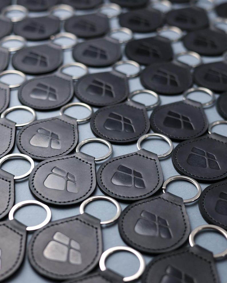 May include: A close-up of a group of black leather keychains with a silver metal ring. Each keychain features a raised, embossed logo of a square with four smaller squares inside.