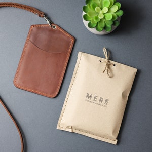 May include: A brown leather ID card holder with a strap and a small pocket. The holder is packaged in a brown paper envelope with the text "MERE FOUNDED IN UKRAINE IN 2012".