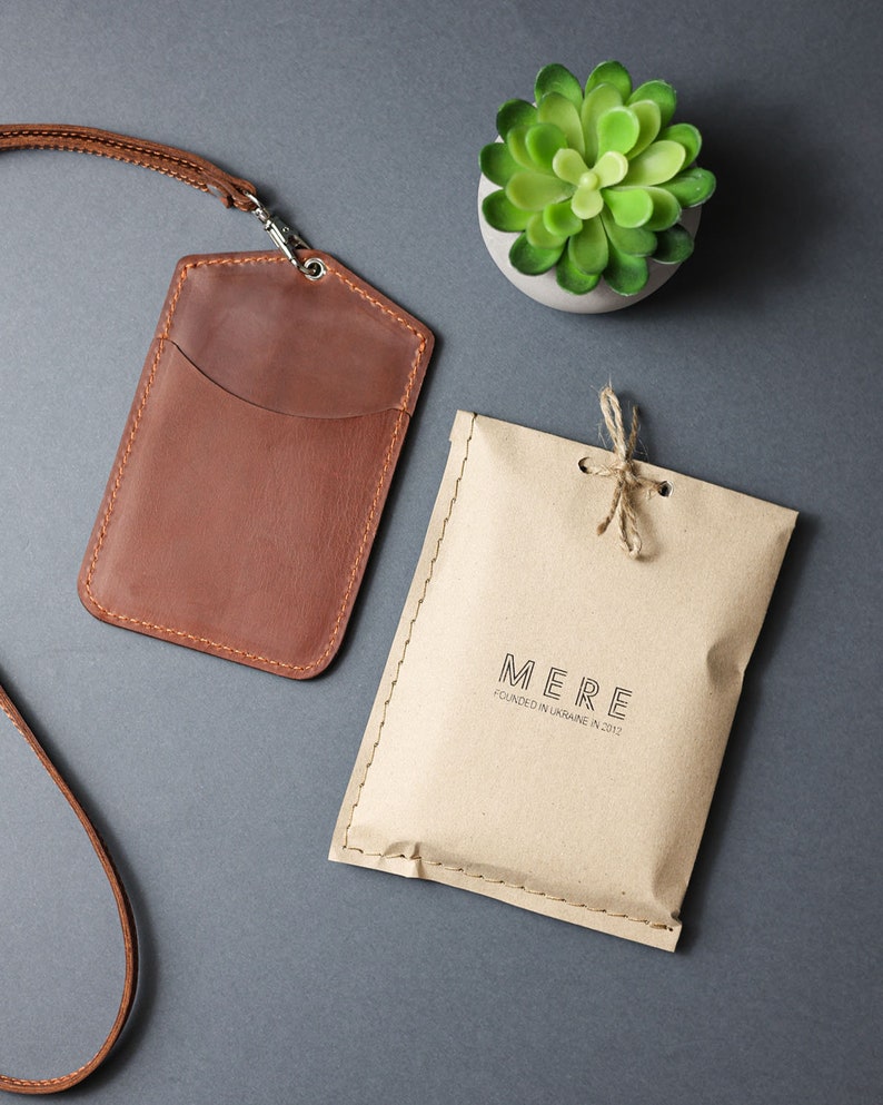 May include: A brown leather ID card holder with a strap and a small pocket. The holder is packaged in a brown paper envelope with the text "MERE FOUNDED IN UKRAINE IN 2012".