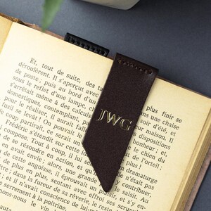 May include: A dark brown leather bookmark with gold initials "JWG" is placed in a book. The bookmark has a pointed end and is made of a smooth material. The book's pages are yellowed, suggesting age. The bookmark is a reading accessory.