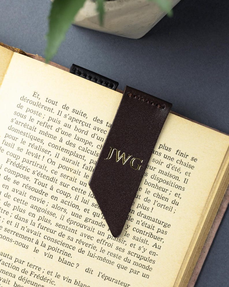 May include: A dark brown leather bookmark with gold initials "JWG" is placed in a book. The bookmark has a pointed end and is made of a smooth material. The book's pages are yellowed, suggesting age. The bookmark is a reading accessory.