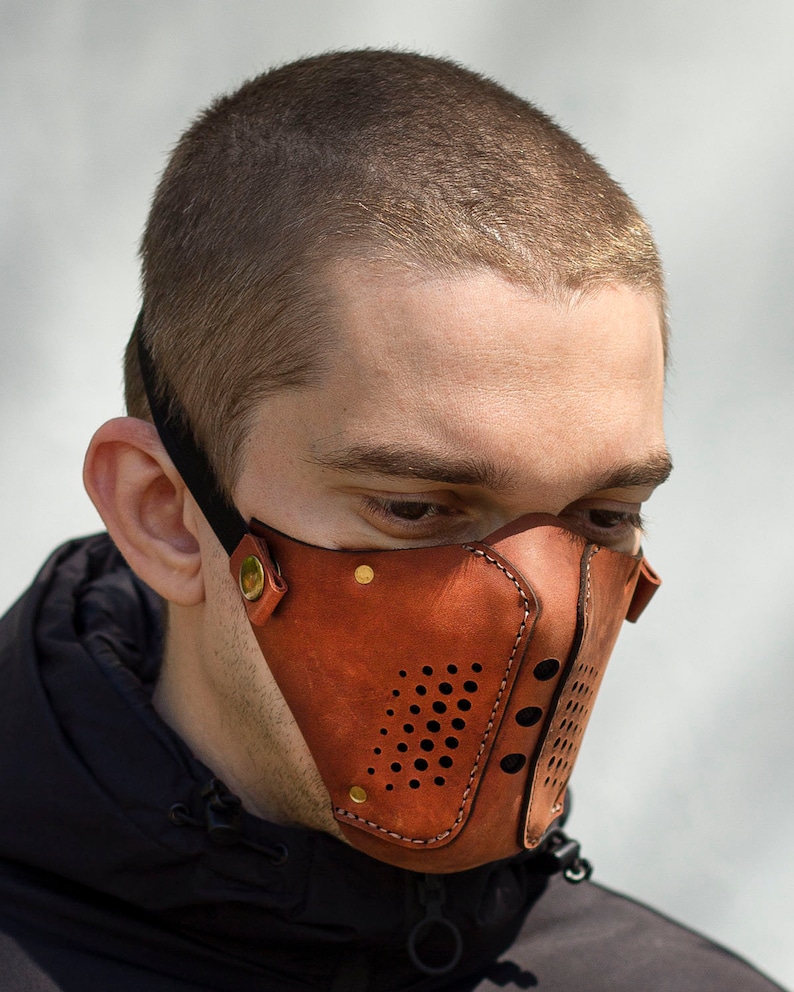 May include: A brown leather face mask with a perforated design and gold accents. The mask has a strap that goes around the head.