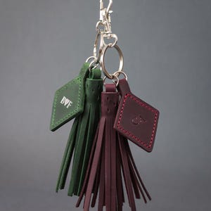 May include: Keyrings with green and burgundy leather tassels. Each keyring has a tag with initials and a silver clasp. The tassels are made of thin leather strips. The keyrings are suspended from a silver clasp.