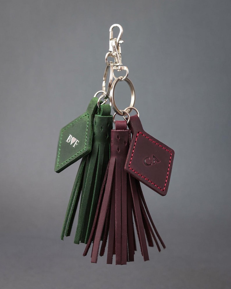 May include: Keyrings with green and burgundy leather tassels. Each keyring has a tag with initials and a silver clasp. The tassels are made of thin leather strips. The keyrings are suspended from a silver clasp.