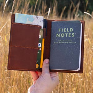 Può includere: Una copertina di diario in pelle marrone con un taccuino Field Notes all'interno. Il taccuino ha una copertina blu scuro con le parole "FIELD NOTES" in bianco. La copertina contiene anche una mappa, carte e una penna. Il diario è aperto.