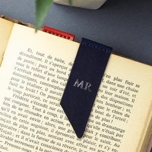 May include: A navy blue leather bookmark with silver initials "MR". The bookmark is in an open book, with a red leather bookmark visible. The book's pages are aged, with French text visible.