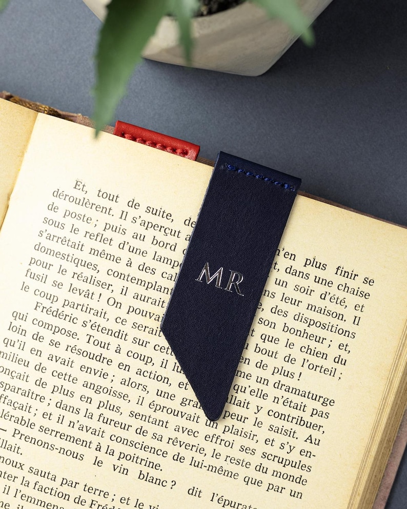 May include: A navy blue leather bookmark with silver initials "MR". The bookmark is in an open book, with a red leather bookmark visible. The book's pages are aged, with French text visible.