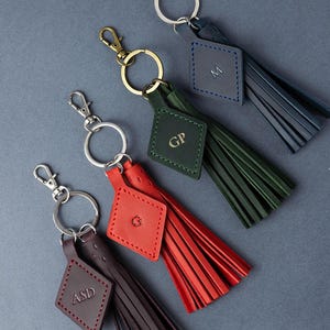 May include: Four leather keyrings with tassels in navy, green, red, and brown. Each keyring has a metal clasp, a ring, and a diamond-shaped tag with initials. The tassels are made of thin strips of leather.