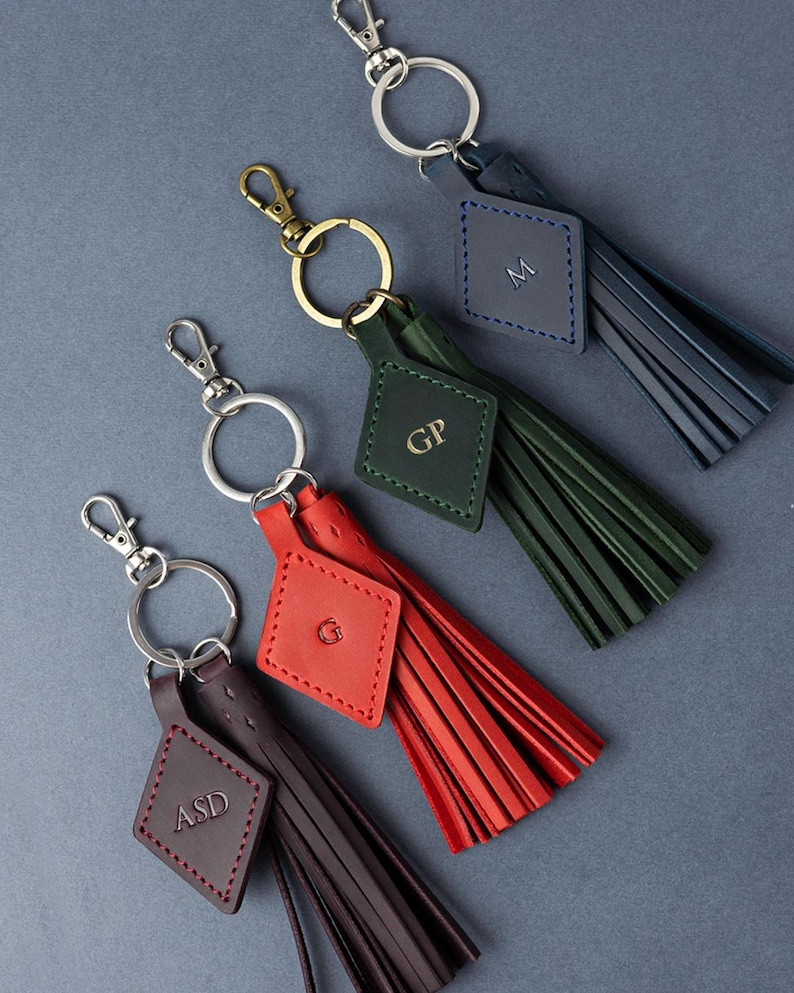 May include: Four leather keyrings with tassels in navy, green, red, and brown. Each keyring has a metal clasp, a ring, and a diamond-shaped tag with initials. The tassels are made of thin strips of leather.