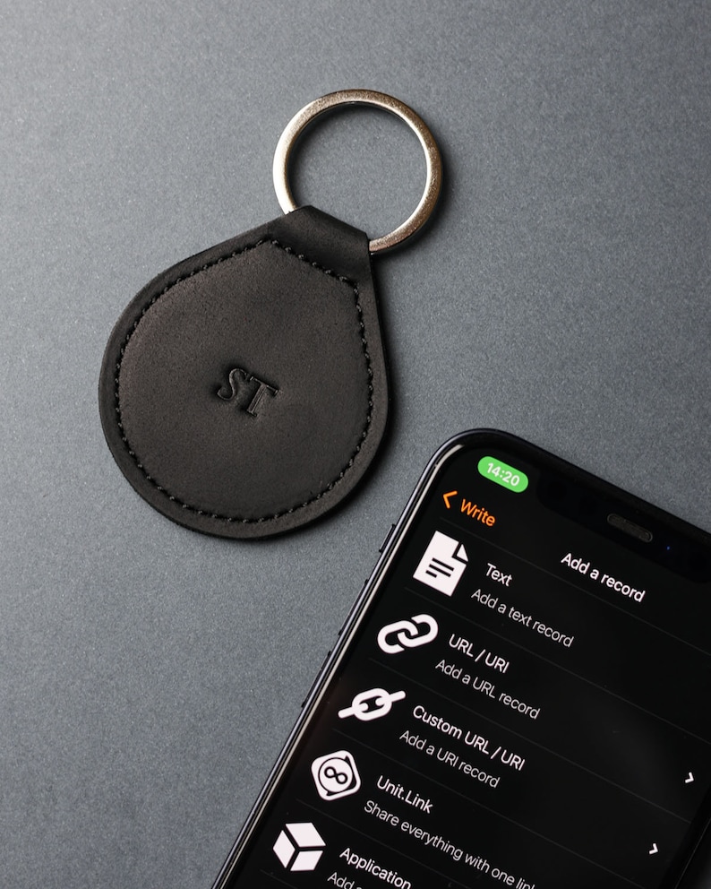 May include: A black leather keychain with a silver ring and the initials "ST" embossed on the front. The keychain is shown next to a smartphone displaying a screen with the text "Write" and a list of options for adding a record, including "Text", "URL / URI", "Custom URL / URI", "Unit.Link", and "Application".