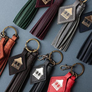 May include: Leather keyrings in a variety of colours, including green, burgundy, grey, brown, black, and red. Each keyring features a tassel and a diamond-shaped tag with a gold or white gazebo design. The keyrings have gold-coloured key rings.
