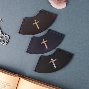 May include: Three leather bookmark corner tabs with a silver cross design. The bookmarks are in brown, dark blue, and black.