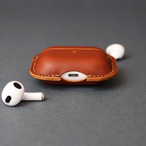 May include: A brown leather protective case for wireless earbuds, featuring visible stitching along the edges and a cutout for a charging port. Two white wireless earbuds with black accents are positioned on either side of the case.