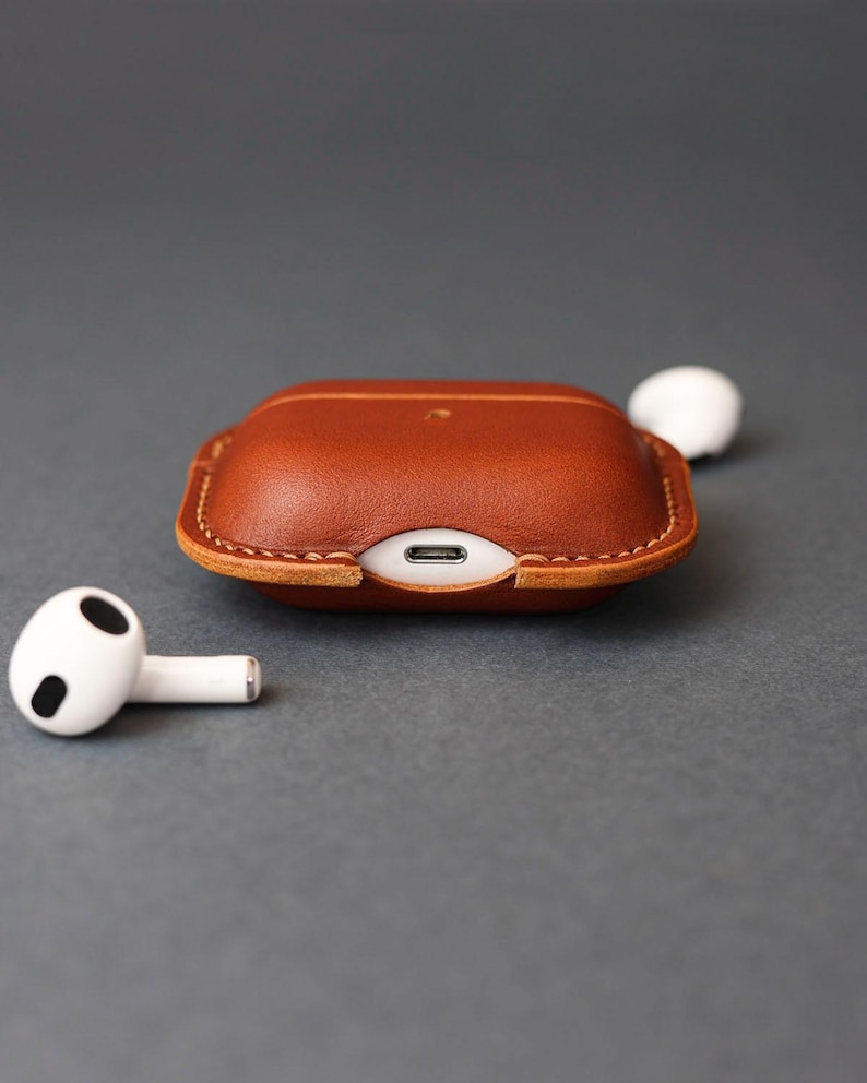 May include: A brown leather protective case for wireless earbuds, featuring visible stitching along the edges and a cutout for a charging port. Two white wireless earbuds with black accents are positioned on either side of the case.