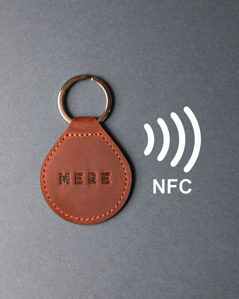 May include: A brown leather keychain with the word "MERE" embossed on it. The keychain has a silver ring and is attached to a small, round, brown leather tag. The tag has an NFC symbol and the word "NFC" printed on it.