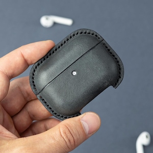 May include: A black leather protective case for wireless earbuds, featuring visible dark stitching along the edges and a small circular cutout on the front surface to reveal the charging indicator light of the enclosed device.