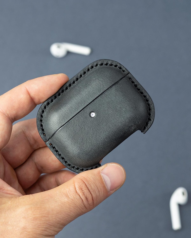 May include: A black leather protective case for wireless earbuds, featuring visible dark stitching along the edges and a small circular cutout on the front surface to reveal the charging indicator light of the enclosed device.