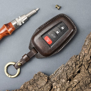 May include: A brown leather key fob with a key chain and a metal ring. The fob has a red button labelled "HOLD" and four other buttons. The fob is lying on a piece of tree bark.