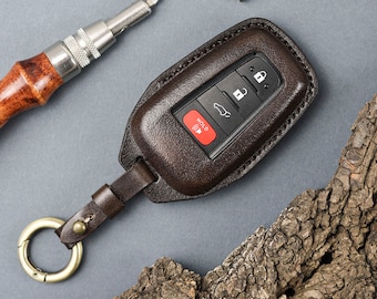 May include: A dark brown leather key fob cover featuring a matching leather strap and a gold-toned metal ring. The cover encases a black car key remote with four buttons: a lock icon, an unlock icon, a trunk icon, and a red button labelled HOLD.