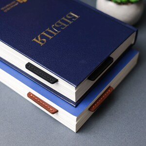 May include: Two blue hardcover books stacked on top of each other. The top book has a gold inscription that reads "Bible". Both books have leather bookmarks.