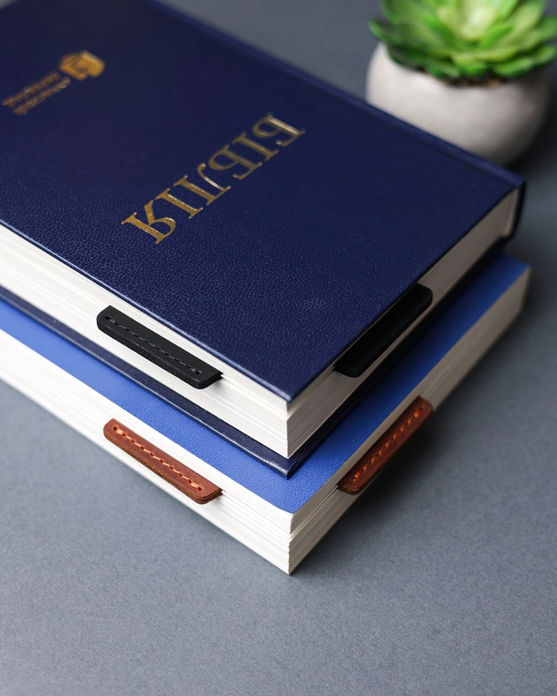 May include: Two blue hardcover books stacked on top of each other. The top book has a gold inscription that reads "Bible". Both books have leather bookmarks.
