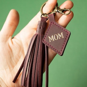 May include: Burgundy leather keychain with a tassel and a tag that reads "MOM" in gold lettering. The tag is diamond-shaped and stitched with a contrasting red thread. The keychain has a bronze-coloured clasp and key ring.