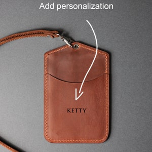 May include: A brown leather ID card holder with a strap. The holder has a pocket for cards and a space for personalization. The text "KETTY" is embossed on the front.