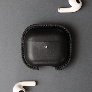 May include: A black leather protective case for wireless earbuds featuring visible stitched edges and a small circular cutout on the front. Two white wireless earbuds are positioned separately above and below the black leather case.