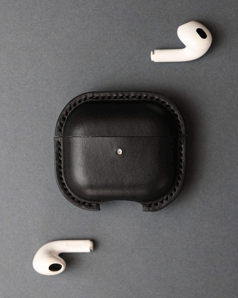 May include: A black leather protective case for wireless earbuds featuring visible stitched edges and a small circular cutout on the front. Two white wireless earbuds are positioned separately above and below the black leather case.