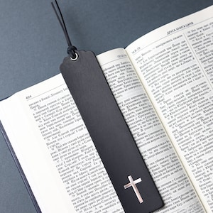 Puede incluir: Marcapáginas de cuero negro con una cruz plateada y un borla negra. El marcapáginas está abierto en una página de la Biblia.