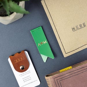 May include: A flat lay image shows a green leather bookmark with gold lettering, a brown leather business card holder, a book, and a kraft paper envelope. The bookmark has the letters "A♡M". The business card holder has the word "MERE" embossed on it.