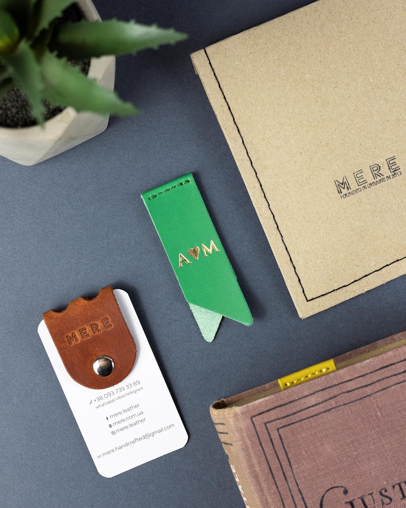 May include: A flat lay image shows a green leather bookmark with gold lettering, a brown leather business card holder, a book, and a kraft paper envelope. The bookmark has the letters "A♡M". The business card holder has the word "MERE" embossed on it.