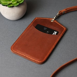 May include: A brown leather ID card holder with a black card inside. The holder has a stitched edge and is attached to a brown leather lanyard.
