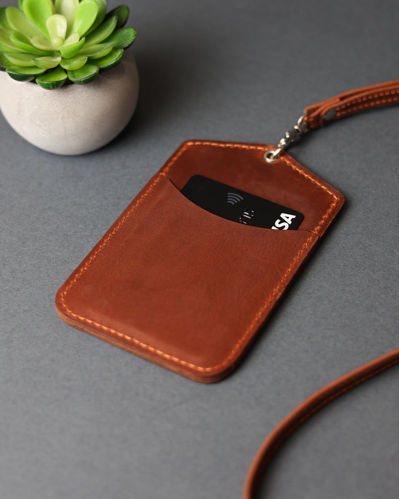 May include: A brown leather ID card holder with a black card inside. The holder has a stitched edge and is attached to a brown leather lanyard.