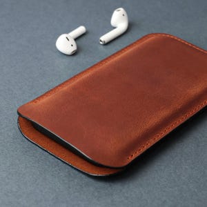 May include: A brown leather case for wireless earbuds. The case is closed and the earbuds are laying next to it.