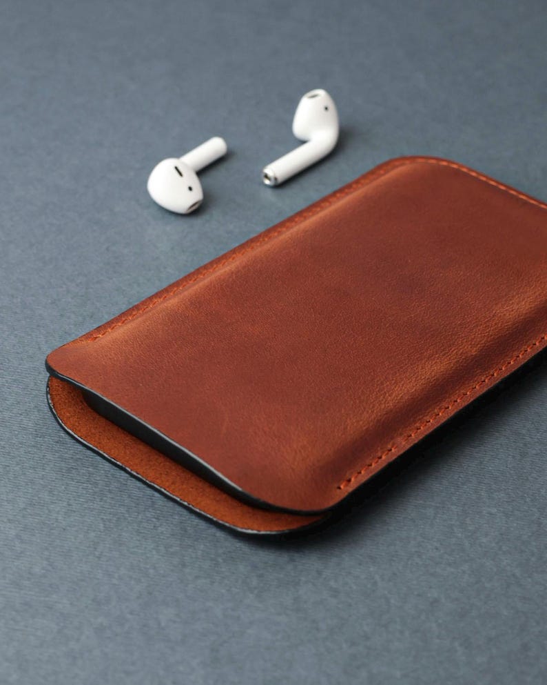 May include: A brown leather case for wireless earbuds. The case is closed and the earbuds are laying next to it.