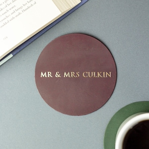 以下が含まれることがあります： 中央に金色のエンボス加工で「MR & MRS CULKIN」と記された、円形のダークバーガンディ色のレザーコースター。コースターは開いた本と、暗い液体が入った白いカップが置かれた緑色のコースターの近くに配置されています。