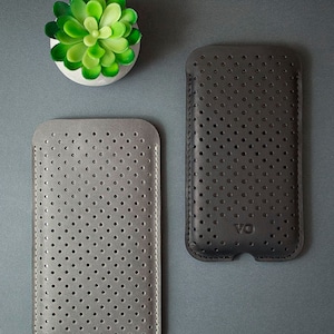 May include: Two perforated leather phone cases, one gray and one dark gray, are displayed on a gray surface. The cases have a stitched border and a series of small, evenly spaced holes. A small, green succulent in a white pot is in the upper left corner.