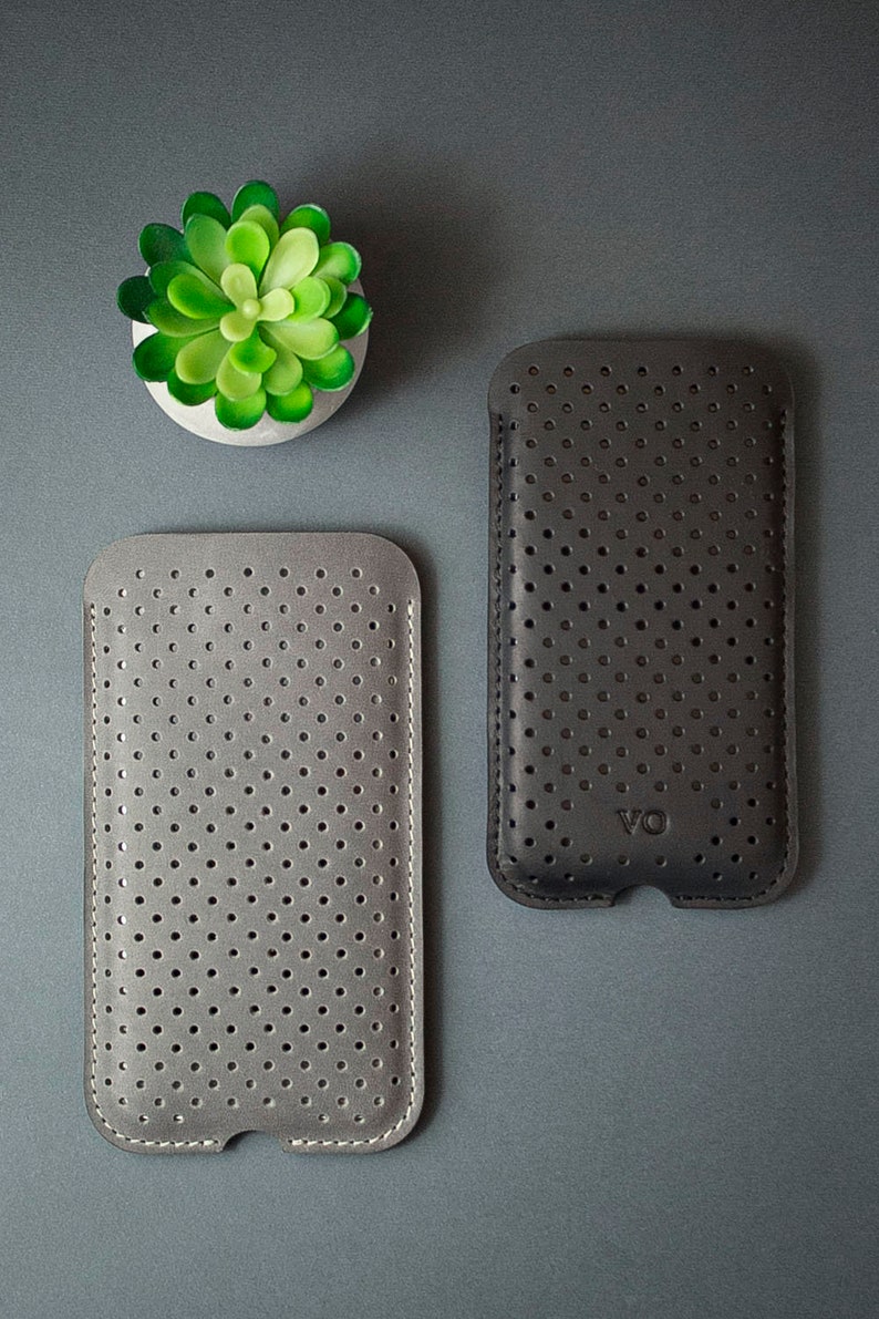May include: Two perforated leather phone cases, one gray and one dark gray, are displayed on a gray surface. The cases have a stitched border and a series of small, evenly spaced holes. A small, green succulent in a white pot is in the upper left corner.